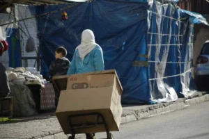 A Palestinian woman pushes a cart with cardboard boxes through a Gaza street lined with makeshift stalls, highlighting the humanitarian crisis and aid shortages.