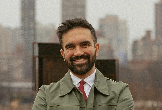 Zohran Mamdani, newly elected mayor of New York City, standing outdoors with city buildings in the background.