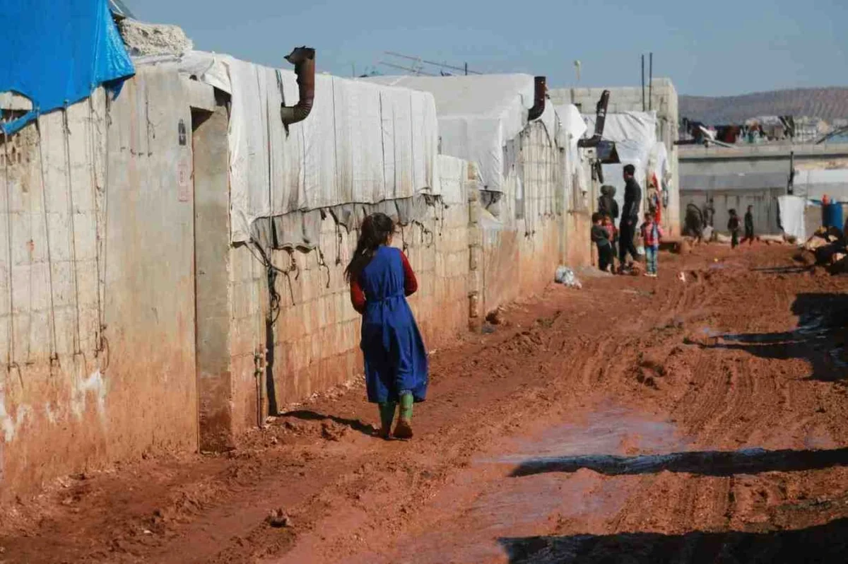 Illustrative image of children walking along a muddy path between temporary shelters, symbolizing displacement and hardship caused by the Syrian war.