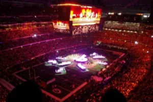 Wide view of a Super Bowl halftime show with a large central stage, giant screens, and a sold-out stadium filled with fans