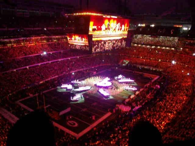 Wide view of a Super Bowl halftime show with a large central stage, giant screens, and a sold-out stadium filled with fans
